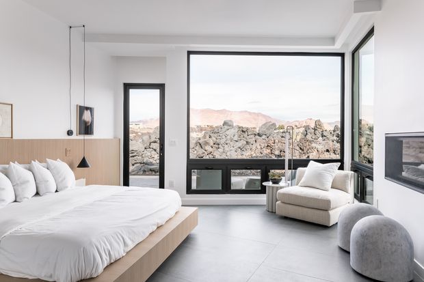 Bright contemporary bedroom with a low light-wood platform bed, white bedding, pale walls, large black-framed windows, a sculptural upholstered lounge chair, and a built-in fireplace. Rocky terrain and mountains are visible beyond the glazing.