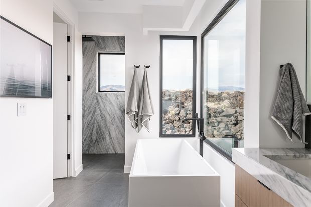 Bright contemporary bathroom with a freestanding white bathtub positioned beside large black-framed windows overlooking a rocky landscape. The room features grey stone-look wall and floor finishes, a marble-look vanity benchtop with timber cabinetry, black tapware, white walls, wall-mounted towels, and a frameless glass shower enclosure visible on the right. A doorway opens to an adjacent marble-look tiled space.