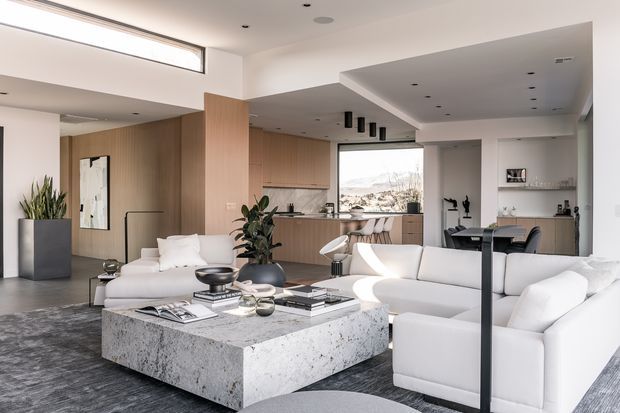 A bright, open-plan contemporary living space with a large white sectional sofa and sculptural armchairs arranged around a substantial speckled stone coffee table. The adjoining kitchen features warm timber cabinetry, a stone island with bar seating, integrated appliances and a large window. White walls, recessed lighting, dark flooring and a dramatic ceiling step create a refined, spacious interior.