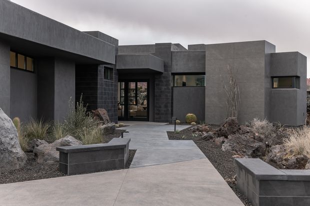 A contemporary low-rise residence with a strongly geometric, flat-roofed form and dark charcoal concrete or rendered masonry walls. The entrance is approached by a broad pale concrete path through a minimalist, xeriscaped garden with gravel, boulders, drought-tolerant grasses, sculptural plants, and built-in dark raised planters. Narrow horizontal windows and recessed glazing reinforce the monolithic modern facade.