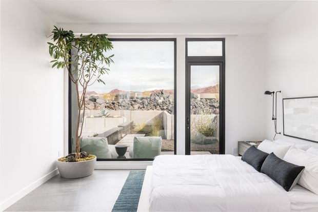 Bright contemporary bedroom with a large bed, white bedding, charcoal accent cushions, a slim black floor lamp, and minimalist artwork. Full-height black-framed glazing opens the room to a small terrace and a rocky landscaped setting, while a tall potted indoor tree adds natural texture.