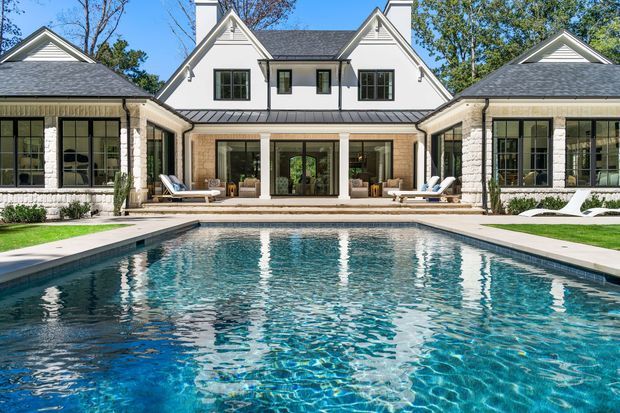 A symmetrical rear exterior view of a large traditional-style home arranged around a rectangular swimming pool. The house has white painted gabled wings, light stone-clad columns and walls, dark grey shingle roofs, black-framed multi-pane windows, and a central glazed living area opening onto a paved pool terrace. Sun loungers and landscaped lawn frame the pool, with mature trees visible behind the building.