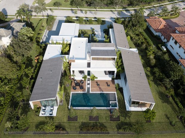 Aerial view of a large contemporary residential property arranged around a central courtyard and rectangular swimming pool. The complex includes a bright white, flat-roofed central pavilion, two long parallel grey-roofed wings, extensive glazing, timber-toned terraces, landscaped lawns, and mature tropical planting. A street and neighbouring homes are visible beyond the property.