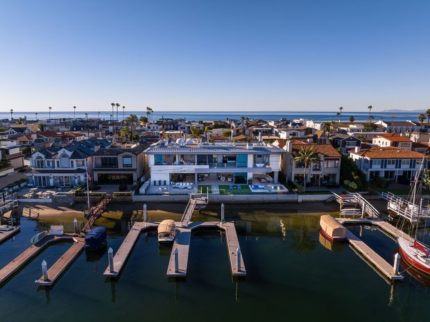 Aerial view of a dense waterfront residential neighbourhood with a prominent modern multi-storey house facing a canal or marina. Private boat docks and moored boats occupy the foreground, while low-rise homes, palm trees, and the open ocean or bay extend into the background under a clear blue sky.