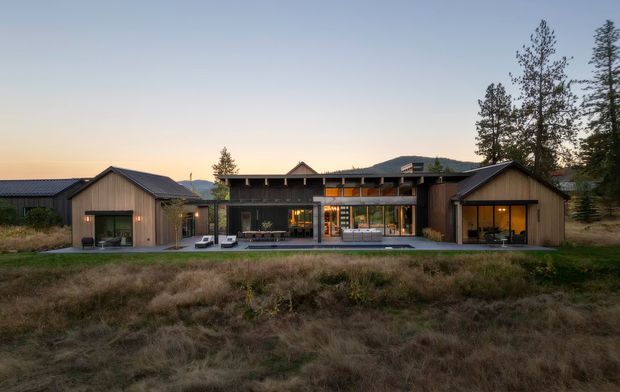 Wide exterior view of a low-rise contemporary residence at dusk, composed of several connected wings with dark timber-clad facades, broad glazing, low-pitched gabled roof forms, and a central glazed living area opening onto a paved terrace. The house sits in a natural grassy landscape with tall trees and distant hills behind it.