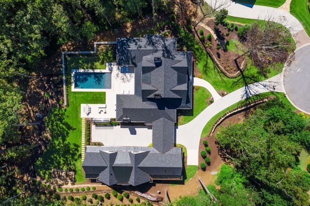 Aerial top-down view of a large residential property surrounded by mature trees and landscaped gardens. The dark grey multi-gabled roof dominates the composition, with visible white-paved terraces, a rectangular swimming pool, curved pathways, and neatly planted garden beds.