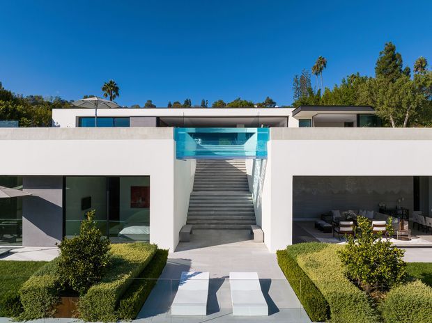 Front-facing exterior view of a minimalist flat-roof residence with strong white geometric volumes, a central open stairway, turquoise glass balustrade or water feature, expansive dark-framed glazing, and formal clipped hedges. The composition emphasizes a symmetrical entry courtyard and clean modern lines under a clear blue sky.