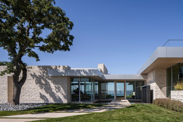 Low-rise contemporary residence with a flat-roofed, rectilinear composition, pale stone masonry walls, extensive dark-framed glazing, and projecting upper volumes. A landscaped lawn and paved path lead to the glazed entry courtyard, with a broad water or coastal horizon visible through the building.