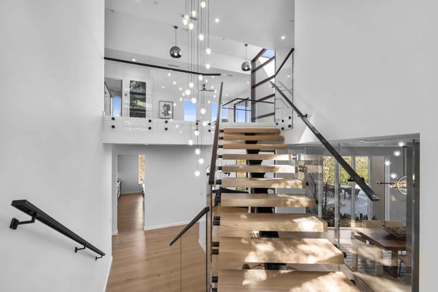 Bright contemporary interior focused on an open floating staircase with thick timber treads, slim dark metal stringers and handrails, and clear glass balustrades. The stair rises through a double-height void with a mezzanine or upper landing, white walls, timber flooring, extensive glazing, and a cluster of pendant globes.