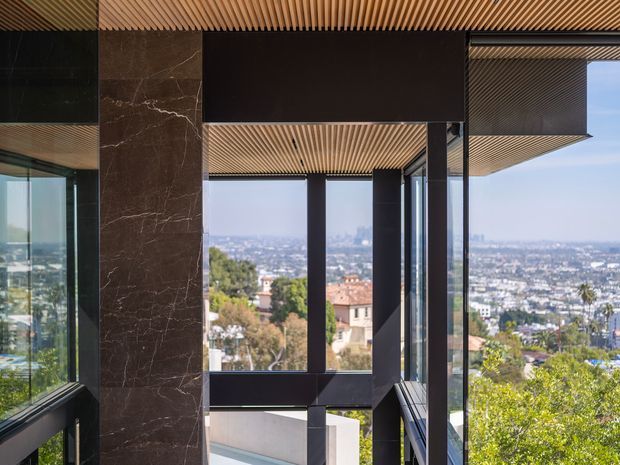 A sophisticated modern interior or covered terrace edge framed by extensive floor-to-ceiling glazing, dark structural columns and a dramatic dark marble-clad vertical wall. A linear timber slatted ceiling adds warmth and texture, while the glazing opens to expansive urban and hillside views. The composition emphasizes strong geometric lines, transparency, and a high-contrast material palette.