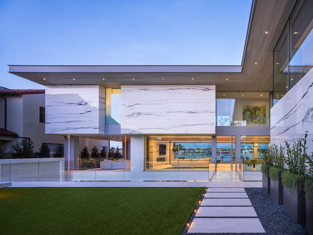 Contemporary multi-level residence viewed from a landscaped front courtyard at dusk. The home features large frameless glazed openings, cantilevered volumes, broad white marble-look wall panels with grey veining, a flat roof with recessed downlights, and a paved stepping-stone walkway across a lawn toward the illuminated entry and living spaces.