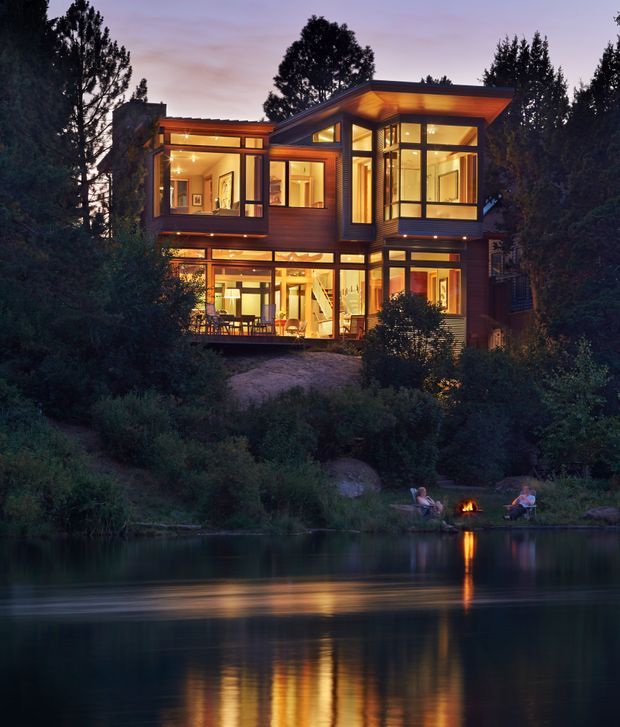 A contemporary multi-storey house set among dense trees beside a calm body of water at dusk. The home has extensive floor-to-ceiling glazing, warm interior lighting, dark timber-toned cladding, projecting flat roof forms, and multiple terraces or balconies. Outdoor chairs and a small fire sit near the water, with the illuminated house reflected in the lake.
