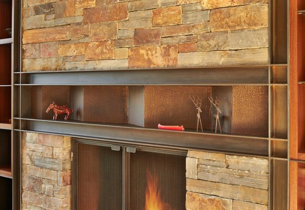 Close-up of a built-in fireplace wall with a rustic stacked-stone surround, dark metal shelving and firebox doors, and warm timber-toned side joinery. Decorative objects are arranged on the open shelves, while a small flame is visible in the lower fireplace.