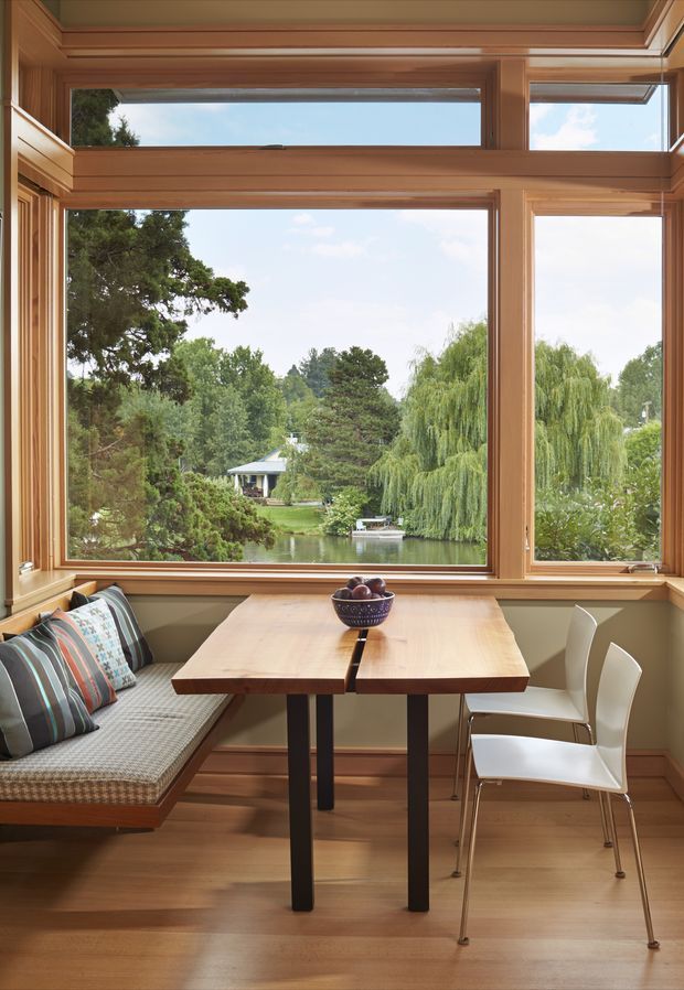 A bright, compact dining nook with a built-in upholstered bench and two white chairs around a simple timber dining table. Extensive warm timber-framed glazing provides views over a landscaped garden and pond. The room has a warm, refined, nature-connected character with built-in window trim and a low, boxed ceiling detail.