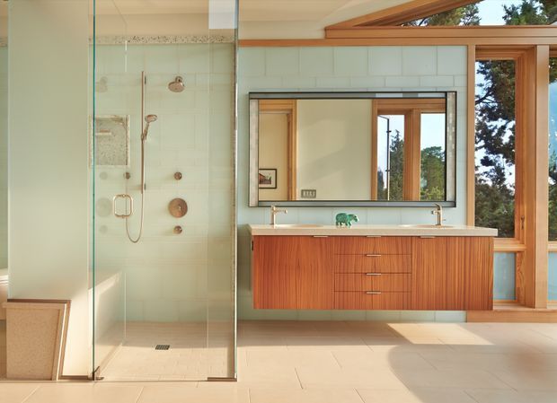 A bright, spacious contemporary bathroom featuring a frameless glass walk-in shower, large wall mirror, floating timber vanity with integrated double basins, and pale tiled flooring and walls. Warm timber-framed windows bring in natural light and views of trees, while subtle decorative tile detailing runs along the shower wall.