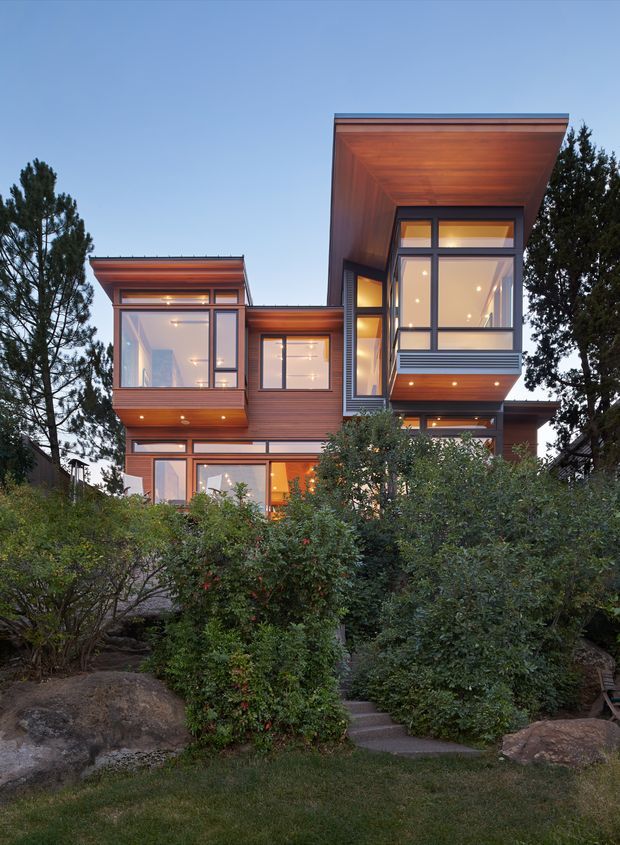 Exterior view of a contemporary multi-level residence set among mature landscaping and rocks. The house features projecting box-like volumes, extensive dark-framed glazing, warm timber cladding and soffits, and subtly sloped or flat roof planes. Evening interior lighting gives the facade a warm glow.