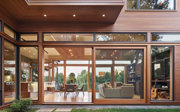 A warm contemporary timber-clad home exterior with extensive floor-to-ceiling glazing. Through the windows, an open-plan dining and living area is visible, featuring timber ceilings, a dining table, sofa seating, built-in shelving, and a guitar. The facade has horizontal timber boards, timber-framed glazing, deep roof overhangs, and recessed exterior lighting, with landscaped planting along the base.
