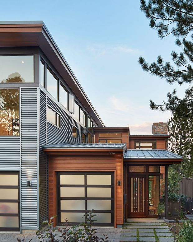 A contemporary multi-storey residential façade with a prominent attached garage, clad in warm horizontal timber boards and contrasting grey corrugated metal panels. Large windows, frosted garage glazing, a covered entry, standing-seam metal roof sections, and a partially visible brick chimney define the composition. Concrete stepping stones lead to the timber-framed front door, with mature trees and planting around the property.