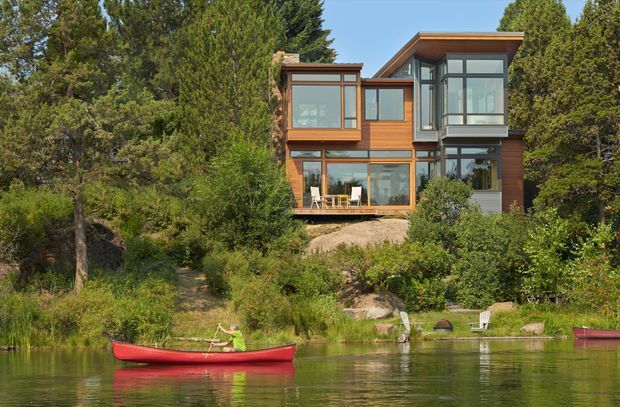 Exterior view of a contemporary multi-level timber-clad house set among dense vegetation on the edge of a calm lake. The home has extensive floor-to-ceiling glazing, projecting box-like rooms, dark window frames, and a prominent sloping roof, with a red canoe on the water in the foreground.