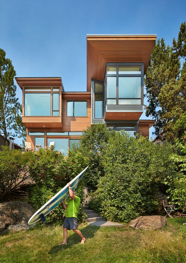 Exterior view of a contemporary multi-level house set among mature trees and dense landscaping. The facade combines warm horizontal timber cladding and deep timber soffits with large blue-grey glazed windows, projecting box-like volumes, and flat or very low-pitched roof planes. A landscaped path and steps lead toward the home; a person carrying a board appears in the foreground.