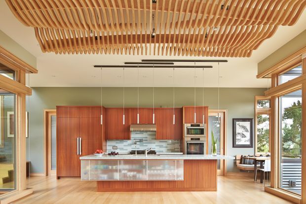 A bright contemporary kitchen with extensive timber cabinetry, a large central island, and a striking sculptural ceiling made from repeated curved timber slats. Full-height timber-framed glazing surrounds the open-plan space, with a dining area visible to the right. The kitchen features integrated appliances, a pale stone benchtop, timber flooring, and muted green walls.