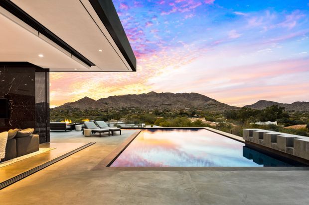 A luxurious contemporary outdoor terrace overlooking a mountain landscape at sunset. The scene features a large rectangular infinity-edge swimming pool, concrete paving, dark stone or marble wall cladding, integrated lounge seating, a linear fire feature, and a broad flat roof with recessed lighting. The pool reflects the vivid pink, blue, and golden sky.