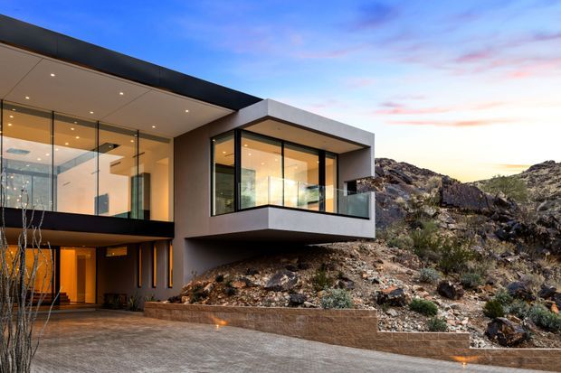Exterior view of a contemporary multi-level home integrated into a rugged rocky hillside. The building features projecting box-like volumes, extensive floor-to-ceiling glazing with dark frames, a flat roof, recessed exterior lighting, and warm-lit interiors visible through the glass. A paved driveway and low retaining wall occupy the foreground, with sparse natural rock landscaping surrounding the structure.