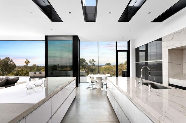 A bright, spacious contemporary kitchen with two large white marble-look islands and extensive white cabinetry. Black-framed floor-to-ceiling glazing opens views toward a coastal or suburban landscape, while a black-and-white marble splashback, dark reflective wall panels, integrated appliances, and dramatic recessed linear ceiling details create a refined high-contrast interior.