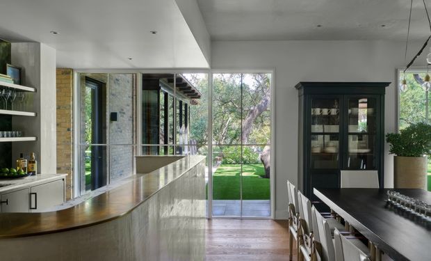 A refined open-plan kitchen and dining area with a long curved island, dark cabinetry, integrated shelving, and large floor-to-ceiling glazing opening toward a leafy garden. The space combines pale stone or plaster surfaces with warm timber flooring, a dark dining table, and abundant natural light.