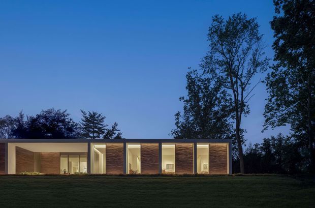 A low, single-storey modern residence photographed at dusk, set within a broad lawn and surrounded by mature trees. The rectilinear facade is composed of large floor-to-ceiling glazed openings, warm illuminated interiors, and repeated vertical bays with dark timber-toned wall panels and slim dark frames. The building has a strong horizontal profile and appears to use a flat roof.