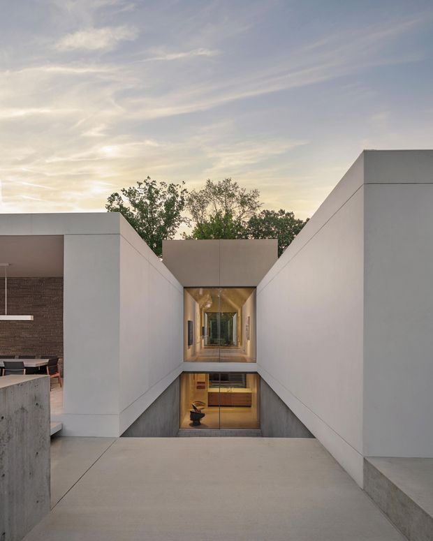 Exterior architectural photograph of a minimalist modern residence featuring crisp white rendered walls, strong geometric planes, a recessed glazed corridor, and a sunken lower-level courtyard or interior space. A concrete terrace leads toward the central entrance, while warm timber surfaces and interior lighting are visible through the glazing. The composition includes an open-air dining area at left and mature trees against a soft evening sky.