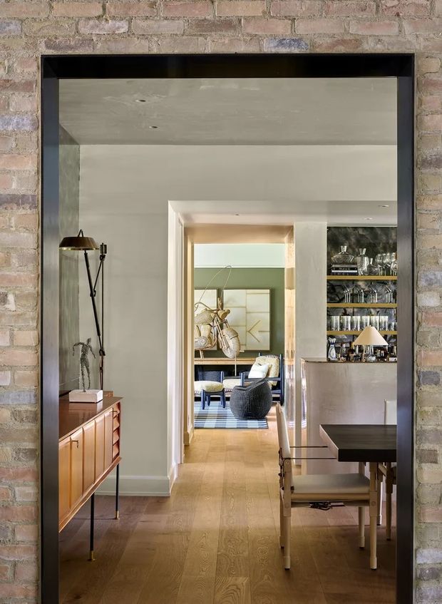 A view through a wide black-framed opening in an exposed brick wall, looking along a warm, refined interior. The foreground contains a timber dining table with upholstered chairs, a timber sideboard, and a floor lamp. Beyond is a compact bar or drinks cabinet with open glassware shelving, leading to a relaxed sitting area with armchairs, artwork, and a patterned rug. Pale plaster walls and ceiling contrast with the exposed brick surround and warm timber flooring.