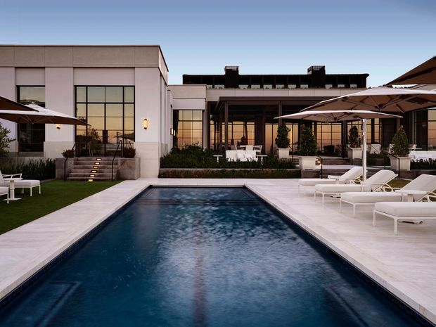 A refined modern courtyard-style exterior centered on a rectangular swimming pool, with pale stone paving, white sun loungers, large umbrellas, manicured planting, and a low-rise building with extensive dark-framed glazing and flat rooflines in the background.