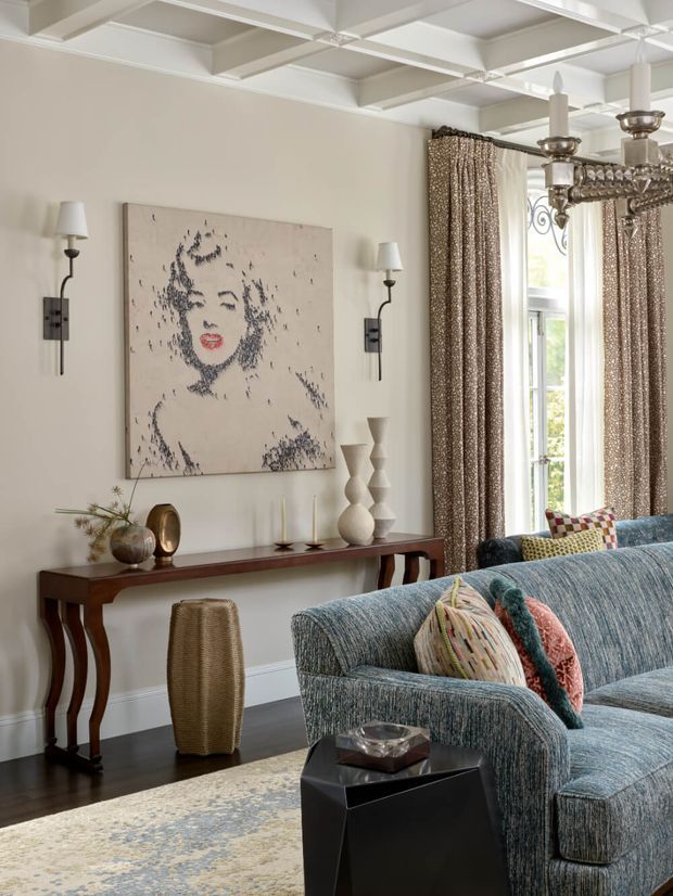 A refined living room with a blue textured sofa, patterned cushions, dark wood console table, sculptural ceramic objects, and a large monochrome portrait. The room features tall patterned curtains, large windows, dark timber flooring, a pale rug, and an ornate silver chandelier. A white coffered ceiling with exposed beam-like detailing adds architectural character.