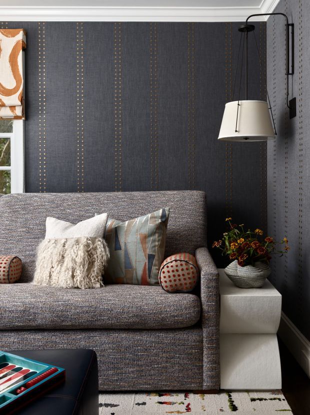 A richly styled living room corner featuring a tailored grey upholstered sofa against dark charcoal patterned wallpaper with vertical dotted stripes. The sofa has textured cream and geometric cushions, a sculptural patterned bolster, and a low dark coffee table with a colorful board or book. A black wall-mounted swing-arm pendant with a cream shade hangs above a white geometric side table holding a textured vase of flowers. White painted trim, a partial window with patterned Roman blind, and a softly patterned rug complete the refined, layered interior.