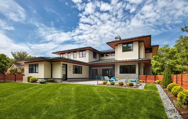 A large contemporary two-storey suburban residence viewed from the landscaped front yard. The home has cream rendered walls, dark-framed windows, broad low-pitched roof planes with deep timber-toned soffits, and a prominent stone-clad fireplace wall beside a covered outdoor entertaining area. A manicured lawn, river-stone drainage strip, timber fencing, and planted borders surround the house.