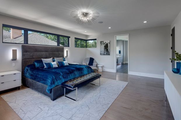 A spacious contemporary bedroom with a large upholstered bed dressed in deep blue bedding, a matching upholstered bench, and a pale geometric rug over warm wood flooring. The room has white walls and ceiling, expansive dark-framed corner windows, minimalist bedside wall lights, a sculptural starburst ceiling fixture, and a partially visible ensuite bathroom through the open doorway.