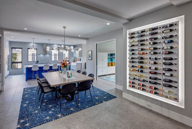 A contemporary open-plan dining and kitchen area with a large rectangular dining table, dark upholstered chairs, a patterned blue rug, and a blue kitchen island in the background. A prominent built-in illuminated wine display occupies the right wall, while a pantry or storage area is visible through an open doorway. The room features recessed ceiling lighting, pendant lights, pale grey walls, and tiled flooring.