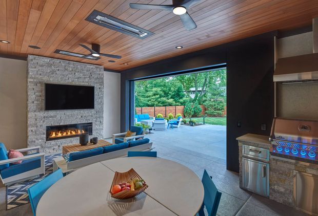 A spacious contemporary indoor-outdoor entertaining room with a timber-lined ceiling, exposed stone fireplace wall, built-in television, ceiling fans, and large retractable glazed opening to a landscaped patio. The room includes a lounge area, round dining table, and a built-in stainless-steel outdoor-style kitchen with grill. The palette combines warm timber, grey stone, dark cabinetry, and vivid blue upholstery.