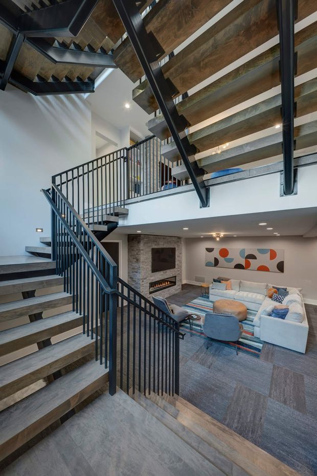 A dramatic multi-level interior centered on an open stairway and mezzanine overlooking a living room. Black metal railings and structural stair supports contrast with timber-toned stair treads and overhead soffits. The lower living area includes a sectional sofa, lounge chairs, a striped area rug, a stone-clad fireplace wall, and abstract artwork. Recessed lighting and the open void create a spacious, contemporary industrial atmosphere.