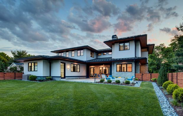 Wide exterior view of a large two-storey contemporary residence with a low-pitched, multi-level roofline, white rendered walls, dark roof and trim, timber-toned cladding accents, extensive windows, and a covered outdoor living area. The house is set behind a broad manicured lawn with landscaped garden beds, timber fencing, and a stone-edged pathway under a dramatic evening sky.