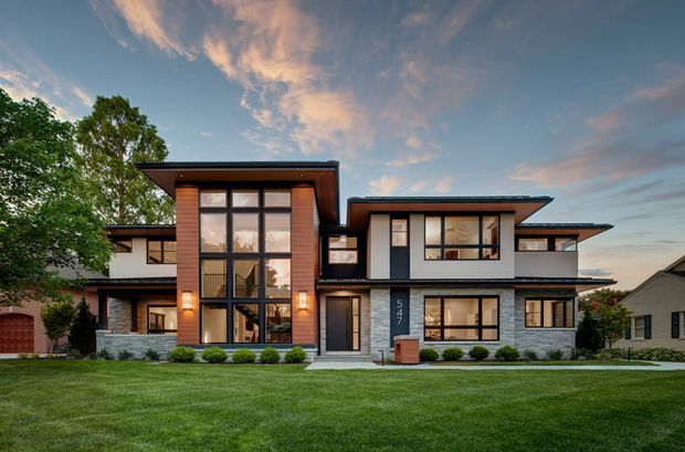 Exterior architectural photograph of a large contemporary multi-level residence viewed from the front across a broad lawn. The home has layered low-pitched, flat-style roof planes with deep overhangs, extensive glazing, dark trim, light rendered walls, grey stone veneer, and warm vertical timber cladding around a tall central entry volume. Mature landscaping and neighbouring homes frame the property under an evening sky.