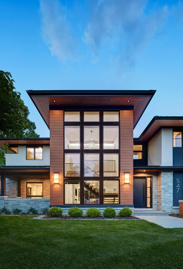 Front exterior view of a large contemporary multi-storey residence. The facade features a dramatic projecting low-pitched roof over a tall central entrance volume, extensive floor-to-ceiling dark-framed glazing, warm horizontal timber cladding, pale stone veneer, and rendered wall panels. A landscaped lawn, clipped shrubs, and pathway occupy the foreground, while the glazed entry reveals a stair and chandelier inside.