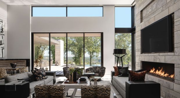 A sophisticated contemporary living room with a double-height volume, expansive floor-to-ceiling glazing, and a large sliding glass opening to an outdoor terrace. The room features a substantial grey stone-clad fireplace wall with a wall-mounted television and linear fire, grey upholstered sofas and ottomans, patterned accent chairs, dark timber details, and sculptural decorative pieces. Mature trees and water are visible beyond the glazing.