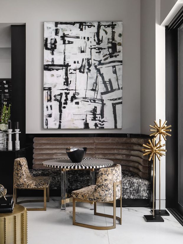 A sophisticated modern sitting area with a black-and-white abstract artwork, dark grey walls, a sculptural ribbed banquette, patterned armchairs, a round monochrome striped table, and distinctive gold starburst floor lamps. The room has a dramatic, high-contrast palette with polished metallic and stone-like details.