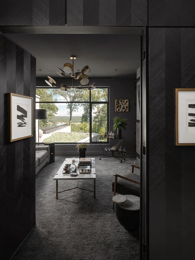 A dark, sophisticated living room viewed through a wide opening, with charcoal chevron wall panelling, matching dark built-in surfaces, a large black-framed picture window, grey carpeting, a light grey sofa, sculptural lounge chairs, and a low rectangular coffee table. Abstract framed artworks and a distinctive multi-disc pendant light add visual interest, while the window opens to a leafy suburban landscape.