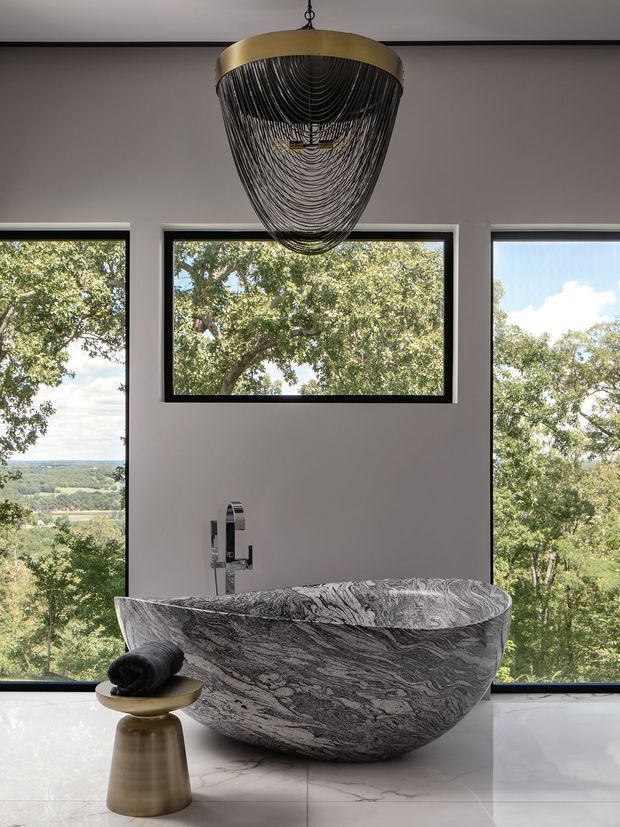 A luxurious contemporary bathroom featuring a sculptural freestanding stone bathtub with dramatic grey and white veining, positioned in front of large black-framed windows overlooking a leafy landscape. A chrome wall-mounted tap, pale walls, light tiled flooring, and a striking gold-and-black pendant light complete the refined space.