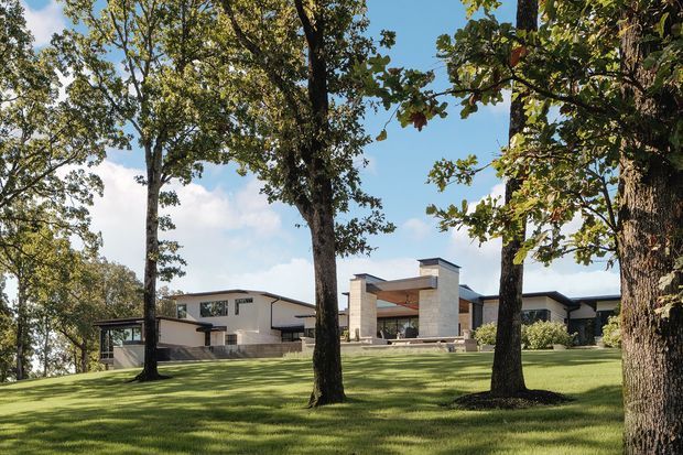 A broad exterior view of a contemporary low-profile residence set within a manicured, gently sloping lawn and mature deciduous trees. The house has pale rendered or masonry walls, dark-framed glazing, strong horizontal rooflines, and a sheltered central outdoor entertaining area framed by tall stone-clad elements. Clear blue sky and extensive established landscaping create an open suburban or rural setting.