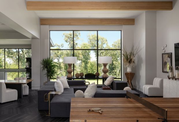 A bright, contemporary living room with a large black-framed, floor-to-ceiling window overlooking trees. The room features dark modular sofas, white upholstered armchairs, a large timber coffee table, sculptural table lamps, indoor planting, and exposed timber ceiling beams. A dark herringbone floor and neutral plaster walls create a refined, modern interior.