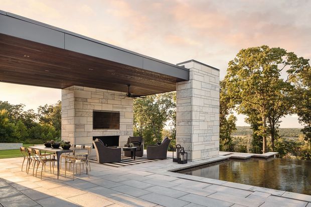 A refined modern outdoor entertaining area with a large dining table and lounge seating beneath a broad cantilevered roof. Pale rectangular stone-clad columns frame a built-in fireplace and television, while a dark timber-lined soffit adds warmth. Large-format pale paving extends around an infinity-edge pool overlooking a wooded landscape.